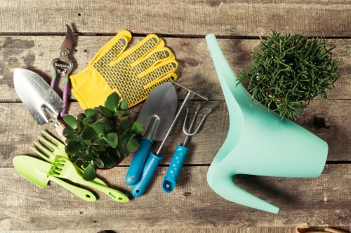 A flat lay image of gardening tools and accessories arranged on a weathered wooden surface, including a pair of yellow gardening gloves, a small hand trowel, a cultivator with purple handles, a hand fork with blue handles, and a bright green hand rake, all positioned around a small, round green potted plant with dense foliage. On the right side of the image, there is a light aqua plastic watering can with a long spout. The scene captures the essentials for gardening and outdoor maintenance, suitable for a garden in Penge, London, emphasizing a well-organized setup for flower bed creation or general garden care by a professional landscapers team like Landscapers Penge.