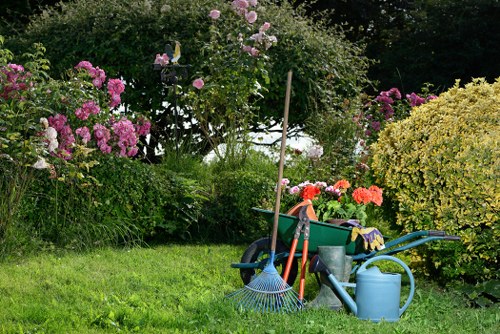 The image depicts a well-maintained garden with a lush, green lawn in the foreground. Surrounding the lawn are various flowering bushes and shrubs, including pink, purple, and yellow blooms, providing vibrant colour contrasts. In the background, there is a mix of taller trees and dense foliage, creating a natural backdrop for the outdoor space. On the right side of the image, gardening tools and supplies are arranged on the grass, including a green wheelbarrow filled with cloths, a blue watering can, a raked broom, and a pruning shear, indicating active garden maintenance. The weather appears clear with bright, natural daylight highlighting the vibrant plant colours and lush textures of the garden. The scene suggests a typical outdoor area in Penge, suitable for professional gardening services such as lawn care, shrub pruning, and general garden upkeep, contributing to a tidy and attractive landscaped environment maintained by experts like Landscapers Penge.