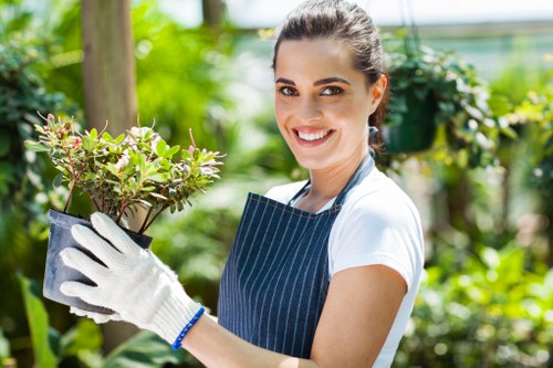 A woman with dark hair tied back, wearing a white t-shirt, a blue striped apron, and white gardening gloves, is standing outdoors in a lush garden. She is smiling and holding a small potted plant with green and purple foliage. The background features various green plants, shrubs, and trees, suggesting a well-maintained landscaped garden. The environment appears bright and sunny, with natural light illuminating the scene, indicative of a warm, clear day. The garden includes elements such as soil beds, leafy plants, and a variety of garden greenery, aligned with professional gardening and landscaping services offered by Landscapers Penge, situated in the Penge area of South London, with postcode likely covering SE20 or nearby districts. This image emphasizes gardening activities, plant care, and outdoor garden maintenance typical of local landscape gardening projects.