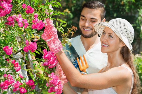 A garden scene featuring a lush, well-maintained lawn with neatly edged flower beds filled with vibrant pink and purple rhododendrons, bordered by a natural soil area. In the background, there are verdant shrubs and mature trees providing shade, with some sunlight filtering through the leaves. The garden appears to be part of a private outdoor space in Penge, with a tidy, landscaped layout that showcases a combination of lawn, flower beds, and natural planting. The overall environment looks bright and inviting, suggesting a mild weather day suitable for outdoor gardening activities. This scene emphasizes the importance of garden maintenance and planting, aligning with services offered by local landscapers in Penge, and highlights the teamwork involved in gardening projects such as planting or pruning.