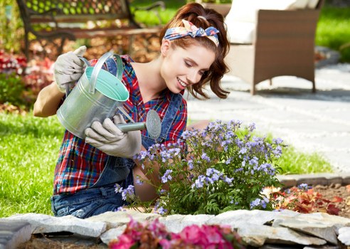 A young woman with light skin, wearing a plaid shirt, gardening gloves, and a headband, is kneeling in a well-maintained garden near Penge, engaging in planting or caring for a small flowering shrub with purple blooms. She is holding a watering can, ready to water the plant, and appears to be smiling while working outdoors on a bright, sunny day. The garden features a low stone border separating the flower bed from the lawn, with lush green grass and other garden plants visible in the background. There is outdoor patio furniture, including a woven chair, placed on a paved area behind her, along with a pathway leading through the garden. The scene reflects a neat, landscaped outdoor space typical of residential gardens that professional gardeners and landscapers, such as those at Landscapers Penge, might maintain, especially in the local area around Penge, SE20 postcode. The overall setting emphasizes healthy plant growth, natural earthy tones, and an inviting outdoor environment suitable for gardening services.