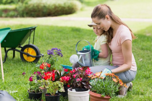 In a well-maintained outdoor garden space, a woman and a young girl are kneeling on a lush, green lawn, engaging in gardening activities. The foreground features a collection of vibrant flowering plants in various pots, including red, purple, and orange blossoms, positioned close together, creating a colourful array against the grass. Behind them, a green wheelbarrow with yellow accents is visible, partially filled with gardening tools and soil, indicating ongoing planting work. The background shows a neatly trimmed hedge and a softly blurred lawn extending into the distance, suggesting a private residential garden in Penge. The scene is lit by natural daylight, under a bright, clear sky, with the woman wearing casual outdoor clothing suitable for gardening, and the girl holding a small gardening trowel, both focused on nurturing their plants. This captured moment highlights gardening activities that a professional landscaper from Landscapers Penge might assist with, including planting, lawn care, and garden maintenance, contributing to a lush, colourful, and well-organised outdoor space tailored to local garden styles and conditions.