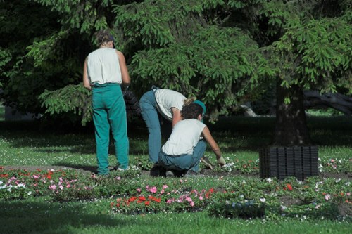 The image depicts three individuals working together in a lush, well-maintained garden in Penge, with dense green foliage and large trees providing a shaded environment. One person stands while two others are crouched down, engaging in planting or gardening activities within a vibrant flower bed bordered by a variety of small, colorful flowering plants in shades of pink, red, and white. The foreground features a neatly trimmed grass lawn, adjacent to the flower bed, and a black garden grid or mat is positioned on the right side, possibly for planting or soil preparation. The backdrop consists of dense, leafy branches, creating a natural canopy that filters soft daylight onto the scene. The environment appears to be part of a landscaped outdoor space, emphasizing the importance of lawn and garden maintenance, likely supported by local gardening services such as those offered by Landscapers Penge, to enhance outdoor beauty and biodiversity in this suburban area.