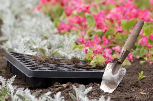 A close-up view of a garden bed in a Penge outdoor space, featuring freshly turned soil with a small gardening trowel inserted into the ground. The soil appears rich and well-drained, ready for planting, with some small weeds or debris visible. In the background, vibrant pink and white flowers border the garden, along with lush green foliage, providing a colorful contrast to the dark soil. The garden layout suggests a neatly maintained yard with areas designated for planting flower beds and decorative shrubs. The scene is illuminated by natural daylight, indicating a clear weather day typical of spring or early summer. The presence of gardening tools and freshly prepared soil illustrates ongoing or upcoming garden renovation or planting work, which Landscapers Penge can assist with, ensuring beautiful, well-maintained outdoor spaces for residents in the local Penge area, within the postcode region.