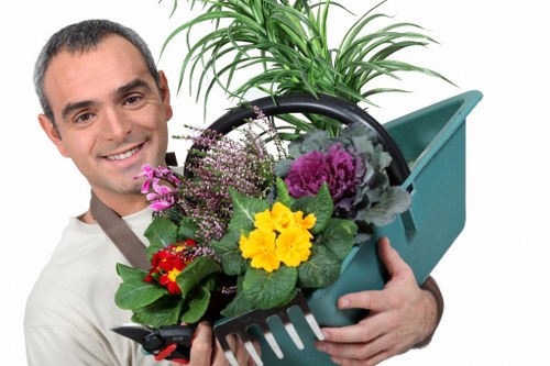 A man with short dark hair and a friendly expression is standing outdoors in a landscaped garden, holding a teal plastic watering can filled with a variety of colorful flowering plants and green foliage. Behind him, neatly maintained grass and a paved patio area are visible, with the garden featuring well-defined flower beds bordered by soil and mulch. In the background, there are trees and shrubs with lush green leaves, suggesting a healthy outdoor environment. The scene is illuminated by natural daylight, indicating clear weather. This setting exemplifies professional gardening and landscape maintenance, aligning with services offered by landscapers in Penge, such as planting, lawn care, and garden enhancement activities.