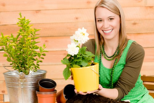 The image shows a smiling woman with shoulder-length blonde hair, wearing a green polka-dot apron over a brown long-sleeve top, seated outdoors in a garden setting. She is holding a yellow flower pot containing a healthy, blooming white flowering plant with green leaves. In front of her on the table are additional gardening items, including a small green shrub in a metallic bucket, a terracotta pot, and an empty black plant pot. The background features a light wooden fence or paneling, suggesting a neatly maintained backyard or patio area within a garden designed for outdoor planting and landscaping. The overall scene emphasizes mature garden planting activity, with lush foliage and vibrant natural tones typical of well-kept outdoor spaces in the Penge area, supporting gardening and landscaping services offered by companies such as Landscapers Penge. The weather appears bright and clear, with natural daylight illuminating the garden area, highlighting the fresh, healthy appearance of the plants and garden tools.