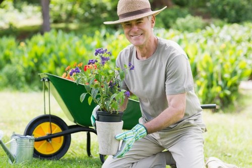 A smiling middle-aged man wearing a straw hat, light grey t-shirt, and gardening gloves kneels on a neatly maintained lawn in a back garden, holding a potted plant with purple and red flowers. Behind him, there is a green wheelbarrow with a bright yellow wheel, partially filled with gardening tools and materials. The garden features a lush, green hedge along the background, with tall trees providing shade and natural backdrop. The lawn is well-kept, with short, dense grass, bordered by flower beds and paved pathways. The weather appears to be sunny and clear, casting natural light across the outdoor space, which exemplifies a well-maintained garden suitable for comprehensive landscaping and lawn care services by companies such as Landscapers Penge.