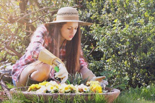 A young woman wearing a wide-brimmed straw hat, plaid shirt, and gardening gloves is kneeling on a grassy lawn in a garden, tending to a flower bed filled with colorful, blooming flowers including yellow, purple, and white varieties. She is using her hands to carefully tend to the plants, with a focused expression on her face. The garden features well-maintained grass, tall hedges, and leafy bushes in the background, creating a lush and vibrant outdoor environment typical of residential gardens in Penge. The scene is illuminated by natural daylight, highlighting the natural tones of green foliage, flower colours, and earthy soil surface, exemplifying outdoor gardening activities with subtle connections to sustainable planting practices. Landscapers Penge often support garden care that includes planting, pruning, and maintaining lush outdoor spaces within the local community.