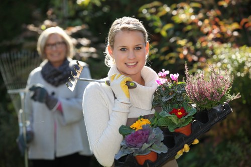 A young woman in a light-colored sweater and gardening gloves is smiling and holding a tray filled with various flowering plants, including purple, yellow, and red blooms, in a well-maintained garden during daylight. In the background, an older woman wearing glasses, a light jacket, and a scarf is slightly blurred, holding a garden tool, with a backdrop of lush greenery, mature trees, and a hedge border. The garden features a neatly mowed lawn, with visible patches of soil near the flower beds, edged by paving or wooden borders, and is bathed in natural sunlight, indicating pleasant weather. The scene reflects outdoor gardening activities typical of residential landscaping in Penge, with vibrant plants and a tidy outdoor space curated for garden care and beautification, aligning with services offered by Landscapers Penge.